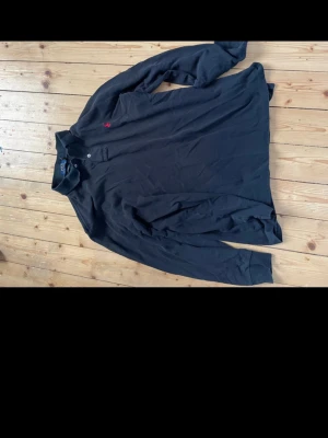 Mörkblå långärmad piké från Polo Ralph Lauren - Klassisk mörkblå långärmad pikétröja från Polo Ralph Lauren med röd broderad logga på bröstet. Skjortan har krage, knappar vid halsen och är tillverkad i mjuk bomull. Perfekt för en clean och stilren look. Finns lite solblekt på kragen se bild. 