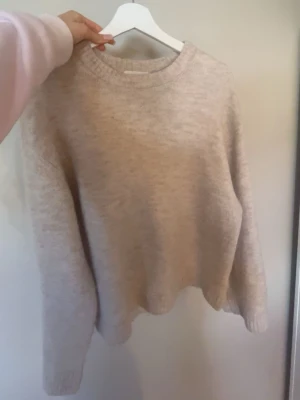 Beige stickad tröja med rund hals - Mysig beige stickad tröja med rund halsringning och långa ärmar. Tröjan har en avslappnad passform och ribbstickade muddar vid ärmslut och nederkant. Perfekt för lager-på-lager och enkel att matcha med jeans eller kjol.