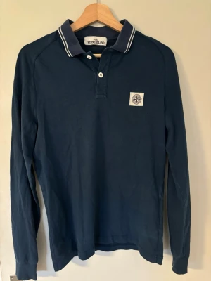 Mörkblå långärmad pike Stone Island - Snygg mörkblå långärmad skjorta från Stone Island med klassisk krage och vit logotyp-patch på bröstet. Skjortan har knappar vid halsen och vita detaljer på kragen. Perfekt för en stilren och avslappnad look.