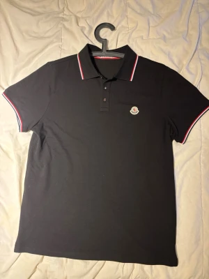 Svart Moncler pikétröja med röd/vit kant - Snygg svart pikétröja från Moncler (Storlek L men funkar på de som har M också) med klassisk krage och knappar framtill. Tröjan har röda och vita detaljer på krage och ärmslut samt Moncler-logga på bröstet. Perfekt för en stilren och sportig look. Jag beställde hem denna via internet och den är tyvärr för liten på mig och vill bli av med den. Den har inte använts då den är för liten på mig och är splitterny. Digitalt kvitto och bevis finns kvar. Prislappen och paketet finns även kvar.