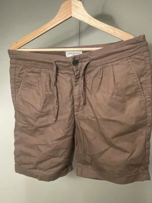 Bruna shorts från John Henric - Snygga bruna shorts från John Henric med dragsko och knapp i midjan. Modellen har två sidofickor och är tillverkad i ett mjukt bomullstyg. Perfekta för en avslappnad och stilren look.