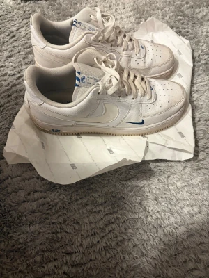 Nike Air Force 1 vita sneakers - Säljer ett par klassiska Nike Air Force 1 sneakers i vitt läder med blå detaljer på loggan och sulan. Skorna har perforerad tå, vit sula och lågt skaft. Perfekta för dig som gillar stilrena och tidlösa sneakers med en twist.