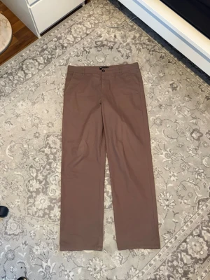 Bruna raka chinos - Snygga bruna chinos med rak passform och klassisk design. Byxorna har framfickor, bälteshällor och stängs med knapp och dragkedja. Perfekta för en clean och stilren look. Materialet ser ut att vara bomull eller bomullsblandning.
