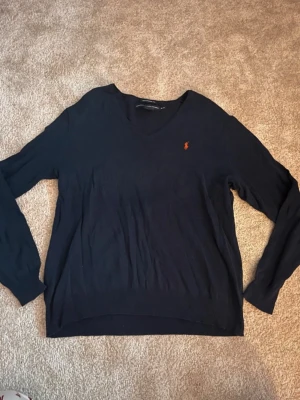 Mörkblå stickad tröja Ralph Lauren - Mörkblå stickad tröja från Ralph Lauren Sport i boyfriend fit. Tröjan har V-ringning, långa ärmar och den klassiska röda loggan broderad på bröstet. Tillverkad i mjuk bomullsblandning för en avslappnad och snygg look.