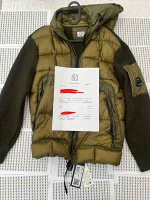 Olivgrön pufferjacka från C.P. Company - Snygg olivgrön pufferjacka från C.P. Company med stickade ärmar och huva. Jackan har dragkedja framtill, två sidofickor med dragkedja samt en ikonisk linsdetalj på ena ärmen. Materialmix av dun och stickad ull ger både värme och stil.