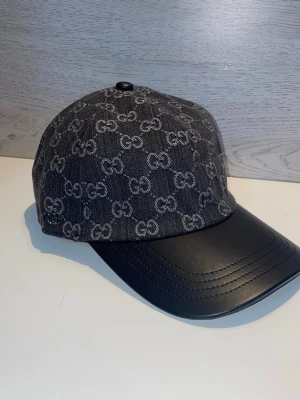 Svart keps från Gucci med GG-mönster - Snygg svart keps från Gucci med klassiskt GG-monogram i grått över hela tyget. Skärmen är i svart läderimitation som ger en exklusiv känsla. Perfekt accessoar för dig som vill ha en trendig och lyxig look.