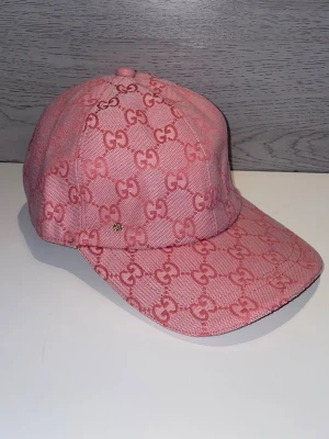 Rosa Gucci keps med GG-mönster - Säljer en snygg rosa keps från Gucci med det klassiska GG-monogrammet över hela kepsen. Kepsen har böjd skärm och är tillverkad i ett slitstarkt tygmaterial. Perfekt för dig som vill sticka ut med en trendig accessoar från ett lyxigt märke.
