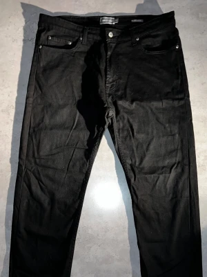 Svarta raka jeans från Dressmann - 38-30 - Säljer ett par svarta raka jeans från H&M med klassisk femficksmodell och knappgylf. Jeansen har en stilren look och är tillverkade i ett mjukt bomullsmaterial med lite stretch för extra komfort. Perfekta till en avslappnad streetstyle.