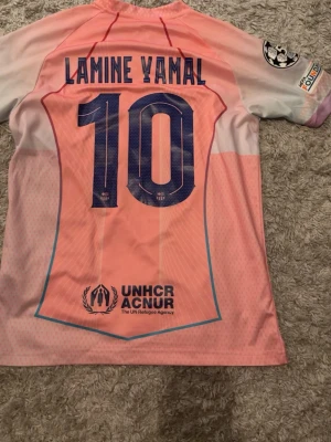 Barcelona Lamine Yamal matchtröja rosa - Säljer en rosa och ljusrosa FC Barcelona matchtröja från Nike med Lamine Yamal och nummer 10 på ryggen. Tröjan har UNHCR-logga, Champions League-märke på ärmen och Spotify-logga på bröstet. Tillverkad i lätt och ventilerande material, perfekt för fotboll.