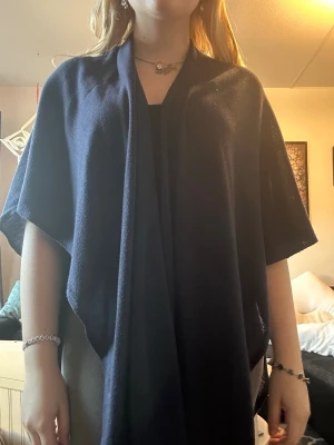 Mörkblå öppen poncho - Mörkblå poncho med öppen front och korta ärmar. Den har en lös och avslappnad passform, perfekt att slänga över en topp eller tröja. Materialet ser ut att vara mjukt och stickat, vilket ger en skön känsla och snyggt fall.