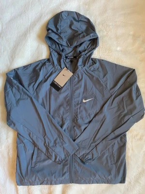Nike grå vindjacka med huva - Snygg grå vindjacka från Nike med huva och dragkedja framtill. Jackan har två sidofickor, elastiska muddar vid ärmsluten och en vit Nike-logga på bröstet. Tillverkad i lätt och tunt material som passar perfekt för blåsiga dagar.