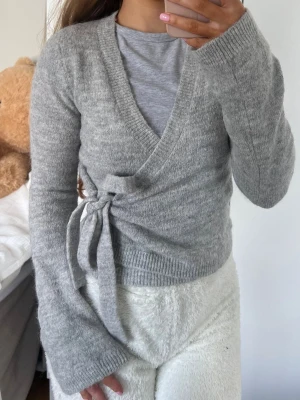 Grå omlottkofta med knytband - Mysig grå kortare omlottkofta med långa ärmar och knytband i sidan. Koftan har ribbade muddar och en mjuk, stickad känsla som passar perfekt till lager på lager. Enkel att matcha med olika outfits för en avslappnad och trendig look. Storlek XS från Gina Tricot 🩶
