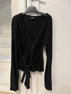 Svart omlottopp från Gina Tricot - Snygg svart omlottopp från Gina Tricot med långa ärmar och knytband i midjan. Djup V-ringning och mjukt, stretchigt material som sitter skönt på kroppen. Perfekt att styla med jeans eller kjol för en trendig look.