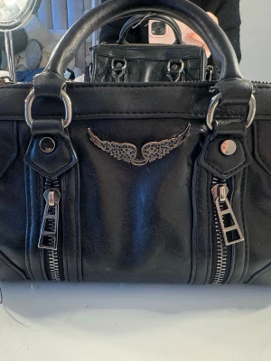 Svart handväska med silverdetaljer - Cool svart handväska i fejkläder med dubbla handtag, två dragkedjor framtill och en snygg silverfärgad vingdetalj. Väskan har en boxig form och metallringar vid handtagen för en edgy look. Perfekt för dig som gillar rockig stil. Lite defekter men inget man tänker på!🖤🖤Likande zadig!!