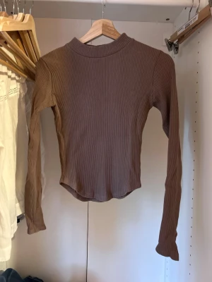 Brun ribbad topp från Gina Tricot - Säljer en brun långärmad ribbad topp från Gina Tricot. Toppen har en högre rundad halsringning och är tillverkad i ett stretchigt, mjukt material som sitter snyggt på kroppen. Perfekt att styla med jeans eller kjol för en trendig look.