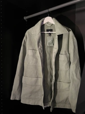 Ljusgrön overshirt från H&M - Stilren ljusgrön overshirt från H&M med fyra stora utanpåfickor och klassisk krage. Overshirten är tillverkad i ett kraftigare bomullstyg och har diskret broderad text på bröstet. Perfekt för lager-på-lager och enkel att matcha.
