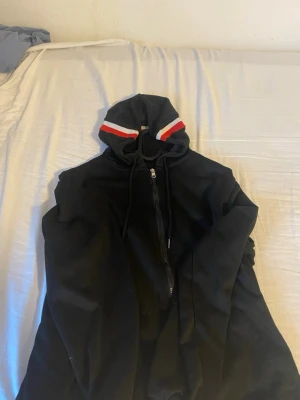 Svart hoodie med rödvit detalj - Säljer en svart hoodie med dragkedja och huva. Huvan har snygga rödvita ränder och samma ränder finns på ärmsluten. Hoodien har dragsko i huvan och är gjord i ett mjukt material, perfekt för chill dagar.