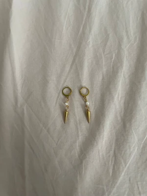 ”esmarilda” örhängen  - Säljer ett par trendiga örhängen i guldfärgad metall med små vita pärlor och konformade hängen. Örhängena har en modern design med ringfäste och passar perfekt för dig som gillar edgy accessoarer med en twist.