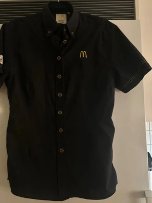 McDonald's svart kortärmad skjorta - Svart kortärmad skjorta med knappar framtill och gul McDonald's-logga broderad på bröstet. Skjortan har klassisk krage och vita kontrastsömmar vid ärmarna. Perfekt för cosplay eller maskerad som McDonald's-anställd.