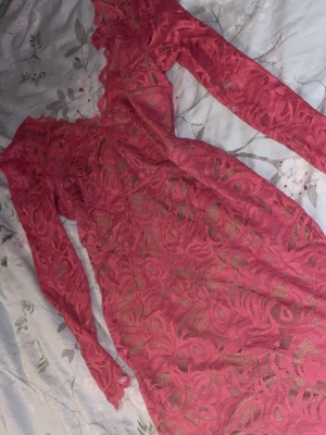 Rosa spetsklänning med lång ärm - Säljer en kort rosa klänning i spets med långärmad design och v-ringning både fram och bak. Klänningen har ett blommigt spetsmönster och är fodrad undertill för extra komfort. Perfekt för dig som vill sticka ut med en feminin och trendig look.