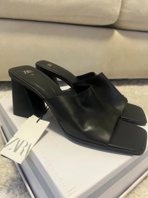 Svarta klackpumps från Zara - Stilrena svarta pumps från Zara med breda remmar och fyrkantig tå. Skorna har en chunky klack och är tillverkade i ett slätt, läderliknande material. Perfekta för dig som gillar minimalistisk och trendig design.