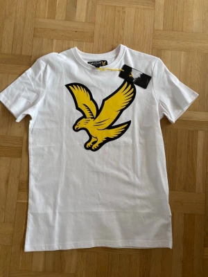 Vit t-shirt från Lyle & Scott - Vit t-shirt från Lyle & Scott med stor gul fågel på bröstet. Klassisk rund hals och korta ärmar. Passar herr Xs