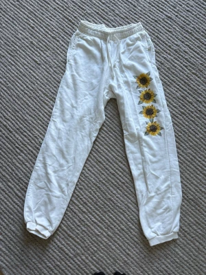 Vita mjukisbyxor med solrosor - Säljer ett par vita mjukisbyxor med solrosmotiv längs ena benet. Byxorna har resår i midjan med dragsko och muddar vid bensluten. Perfekta för chill dagar hemma eller när du vill ha en avslappnad stil.