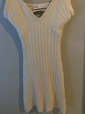 Ribbad vit kortklänning från Bershka - Supersnygg ribbad vit kortklänning från Bershka med v-ringning och figurnära passform. Klänningen är ärmlös och har en stretchig känsla som framhäver siluetten. Perfekt för dig som gillar en enkel men trendig look. Den är även öppne ryggen. Jag har aldrig använt den här klänningen eftersom jag köpte den på vintern så helt oanvänd 