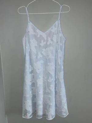 Ljusblå klänning  - Säljer en ljusblå klänning med tunna axelband och v-ringning. Klänningen har ett vackert blommigt spetsmönster och är lätt genomskinligt. Materialet känns mjukt och luftigt.. Knytband i ryggen för justerbar passform.
