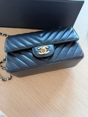 Chanel classic mini flap väska - Chanel Classic Mini Flap Chevron – Svart Lambskin,  light gold hardware     Chanel Classic Mini Flap i svart lambskin med chevron-mönster (V-quilt) och light gold hardware. En elegant och mer unik variant av den klassiska modellen.  Väskan är i bra skick, sparsamt använd. Lätt slitage finns på baksidan vid fickan, vilket är vanligt för lambskin, men inget som påverkar helhetsintrycket.  Full set ingår: Originalbox Dustbag Kvittot från butik Camellia och band + påse   Endast seriösa köpare. Swish