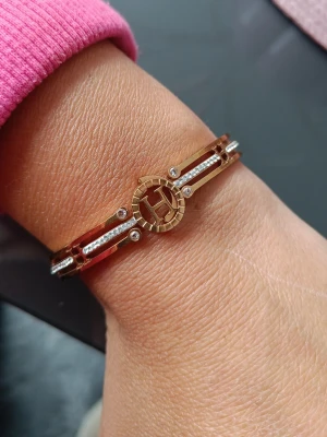 Guldigt armband med H-detalj - Nytt Snyggt armband i guldfärgad metall med gnistrande stenar och en rund H-detalj i mitten. Armbandet har en öppen design med dubbla band och små klara stenar längs sidorna för extra bling. 6 cm diameter. Rostfritt stål och nickelfritt 