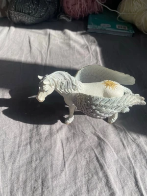 Vit Pegasus-figur i plast - Säljer en detaljerad Pegasus-figur i vit plast med stora vingar och guldfärgad stjärndekor på ryggen. Perfekt som dekoration eller leksak för dig som gillar magiska varelser och fantasy.