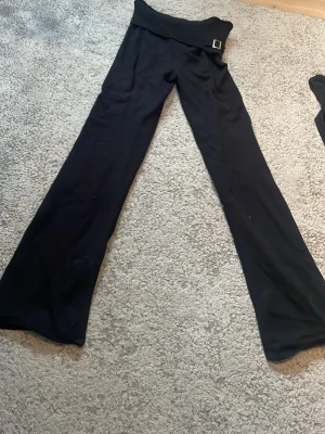 Svarta lågmidjade mjukisbyxor med bred mudd - Säljer ett par svarta mjukisbyxor med bred ribbad mudd i midjan och dekorativt bälte med metallspänne. Byxorna har utsvängda ben och är gjorda i mjukt, stretchigt material som känns skönt mot huden. Perfekta för en avslappnad och chill stil. Ser lite noppriga ut på bilderna men det är de verkligen inte annars de är så fint skick och nästan bara använda några få gånger då de inte riktigt passade mig🥰 skulle säga att de är xs- s i storleken något korta på mig som är 170 