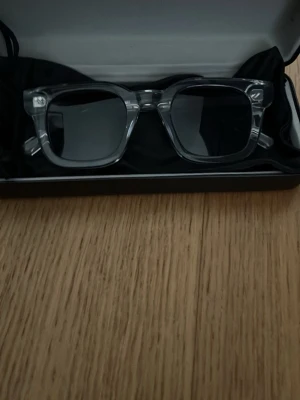 Transparenta solglasögon All Cap One - Snygga solglasögon från All Cap One med transparent, fyrkantig båge och mörka glas. Kommer med svart fodral och putsduk. Perfekta för dig som vill ha en clean och modern look i sommar.