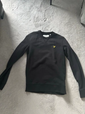 Svart sweatshirt från Lyle & Scott - Svart sweatshirt från Lyle & Scott med rund hals och lång ärm. Tröjan har ett litet gult logomärke på bröstet och är tillverkad i ekologisk bomull. Perfekt för en avslappnad och stilren look.