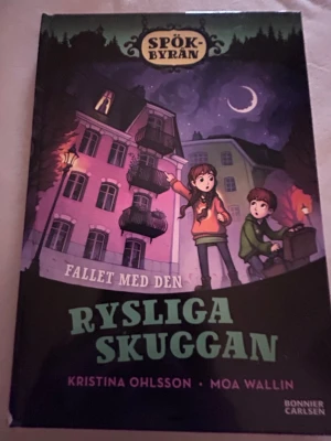 Spökbyrån: Rysliga skuggan - En spännande och klurig bok från serien Spökbyrån, skriven av Kristina Ohlsson och illustrerad av Moa Wallin. Boken har ett mystiskt tema med karaktärerna Kalle och Elsa som löser gåtor om spöken. Omslaget är mörkt med lila och svart, och boken är i pappersformat.Det är en liten repa på boken.