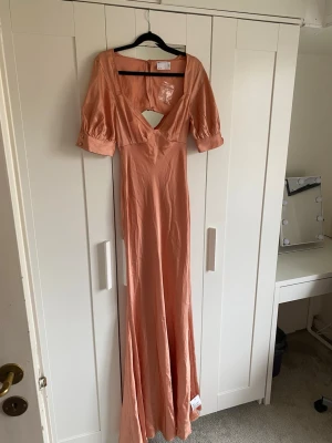 Lång persikofärgad klänning med puffärm - Snygg långklänning i persikofärg med v-ringning och cut-out detalj framtill. Klänningen har puffärmar med resår vid ärmslut och är tillverkad i ett mjukt, glansigt material som ger en elegant look. Perfekt för sommarens alla tillfällen.