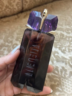 Alien parfym från Mugler  - Alien parfym från Mugler i en unik, mörklila glasflaska med geometrisk form och guldfärgade detaljer på korken. Flaskan har en futuristisk design och Alien-loggan ingraverad på framsidan. Perfekt för dig som gillar ikoniska och snygga parfymflaskor.