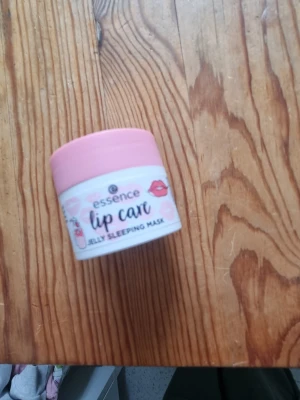 Essence Lip Care Jelly Sleeping Mask - Essence Lip Care Jelly Sleeping Mask i en söt liten burk med rosa lock. Perfekt för att återfukta läpparna under natten. Burken är gjord av plast och har en vit etikett med rosa detaljer och läppmotiv.