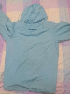 Ljusblå vindjacka med huva - Snygg ljusblå vindjacka med dragkedja framtill och praktisk huva. Jackan har två sidofickor med dragkedja och är tillverkad i ett lätt, vindtätt material. Perfekt för blåsiga dagar och enkel att packa ner i väskan.