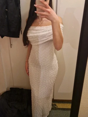 Vit axelbandslös maxiklänning - Säljer en elegant vit maxiklänning med off shoulder-design och figurnära passform. Klänningen är täckt av små pärlliknande detaljer som ger en lyxig känsla. Perfekt för dig som vill sticka ut med en unik och stilren look.
