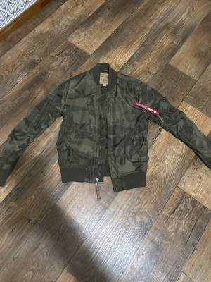 Alpha jacka  - Snygg grön bomberjacka i camouflage från Alpha Industries. Klassisk modell med ribbade muddar, dragkedja framtill och fickor på sidorna. Detaljerad med röd tag på ärmen och utility-ficka. Perfekt för en streetwear-look.