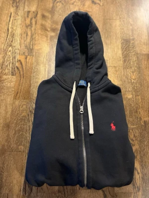 Svart hoodie från Polo Ralph Lauren - Svart hoodie med dragkedja från Polo Ralph Lauren. Klassisk design med vit dragsko i huvan och röd broderad logga på bröstet. Tillverkad i mjukt sweatshirtmaterial, perfekt för en avslappnad stil. Nästan inte använd. 