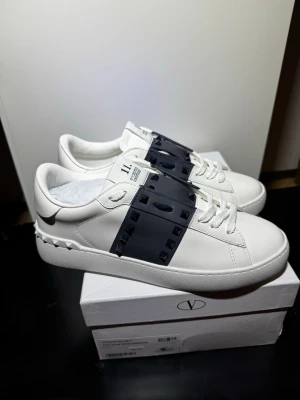 Valentino Rockstud Untitled sneakers - Säljer ett par vita Valentino Rockstud Untitled sneakers med marinblå detaljer och nitar. Skorna har klassisk snörning och är tillverkade i skinn med en cool design och tydlig logga på plösen. Perfekta för dig som vill sticka ut med stilrena men unika sneakers.