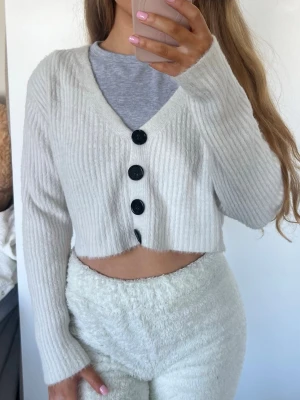 Vit croppad ribbad kofta - Säljer en vit croppad kofta med ribbad struktur och fyra stora svarta knappar framtill. Koftan har v-ringning och långa ärmar, perfekt för en chill och trendig look. Råkat tvätta den med mörka kläder därav går den mot det grå hållet. Storlek S 🩷