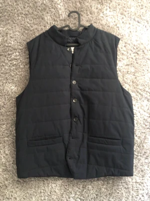 Svart Väst - Stilren svart dunväst från Murrey med ståkrage och knäppning framtill. Västen har två fickor och quiltad design som ger en modern look. Perfekt för lager-på-lager och håller dig varm under kyliga dagar.