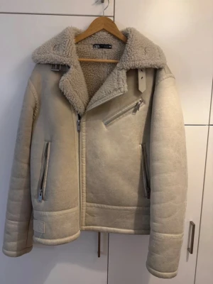 Beige pälsjacka från Zara - Säljer en beige pälsjacka från Zara med mjukt teddyfoder och bred krage. Jackan har dragkedja i sned modell, två framfickor med dragkedja och snygga spänndetaljer vid kragen. Perfekt för kalla dagar och riktigt trendig look.