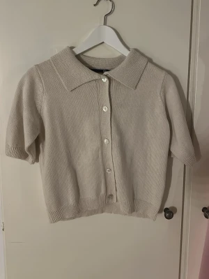 Beige stickad kortärmad kofta 157 - Säljer en beige stickad kofta från 157 med korta ärmar. Koftan har knappar framtill och ribbade muddar vid ärmslut och nederkant. Perfekt att styla över ett linne eller t-shirt. Aldrig använt. 