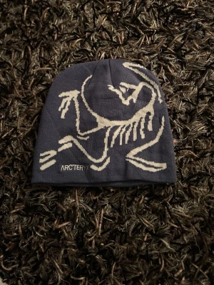 Mörkblå mössa från Arc'teryx - Säljer en mörkblå mössa från Arc'teryx med ett stort vitt fossiltryck på framsidan. Mössan är stickad i mjukt material och har en klassisk, rundad form. Perfekt för kalla dagar och snygg till streetwear-stilen.