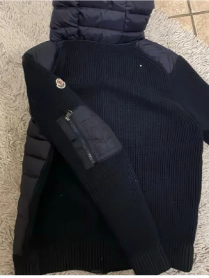 Svart pufferjacka från Moncler - Snygg svart pufferjacka från Moncler med quiltad front och huva. Jackan har ribbstickade ärmar och sidor i ullmix, dragkedja framtill och fickor med tryckknappar. Moncler-logga på ärmen och cool detaljficka på armen . Storlek 2 =M